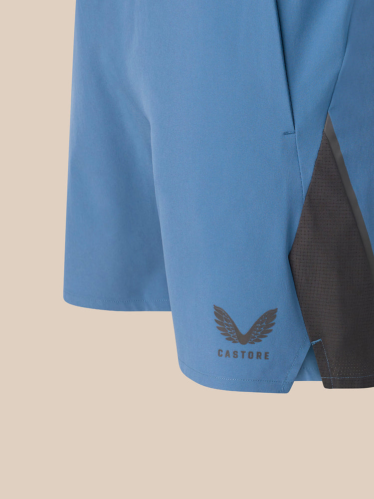 Close-up of a light blue athletic shorts leg with a dark gray mesh side panel and a dark gray Castore wing logo near the hem, set against a solid beige background.