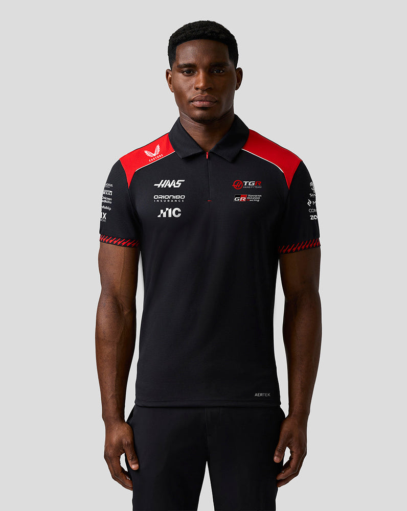 Person wearing a black and red polo shirt with various logos on a gray background