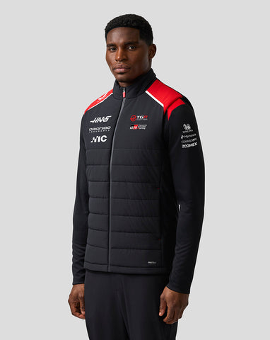 Man wearing a black jacket with various logos on a gray background