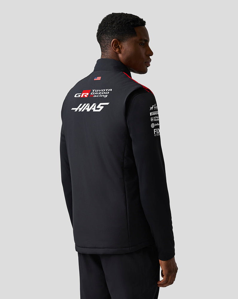 Person wearing a black jacket with sponsor logos on a gray background